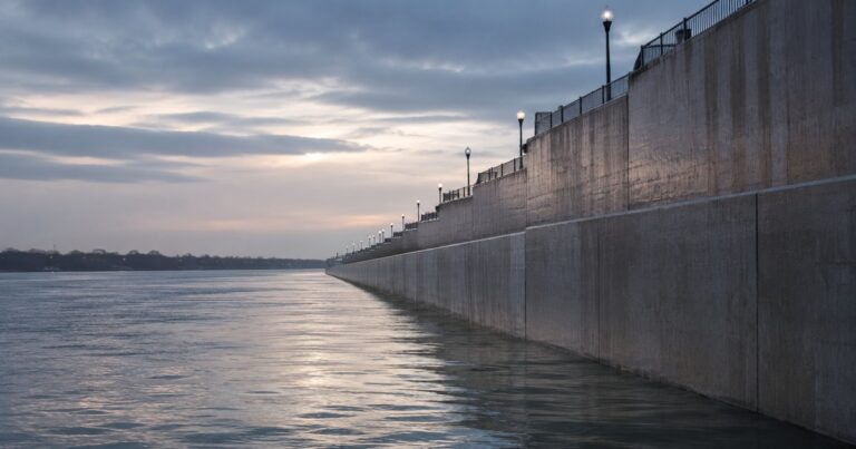 A long reinforced concrete flood-control levee running along a swollen gray-blue Midwestern river at dusk, the water risen partway up the wall with a clear margin of wall height remaining above the waterline and the wall stepping higher as it recedes, no people present — a visual metaphor for how much commercial umbrella limit should sit above a business's expected liability loss.