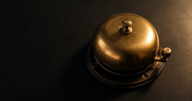A warm-brass bell from a vintage telephone resting on a matte-black surface, illustrating that life events should trigger a phone call to your insurance agent — not a calendar reminder for renewal.
