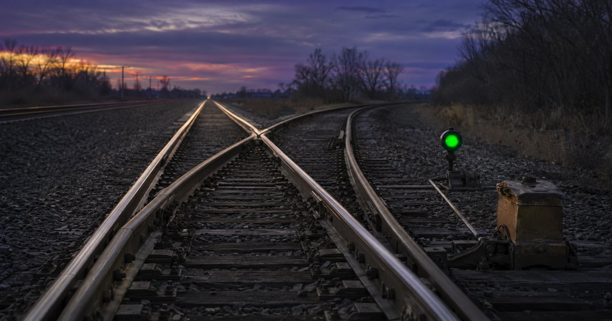A railroad turnout at dusk with the switch points thrown toward a diverging track, a ground-throw switch stand beside the rails, and a dwarf signal glowing steady green — a visual metaphor for a legitimate payment quietly rerouted to the wrong destination while every indicator reads normal.