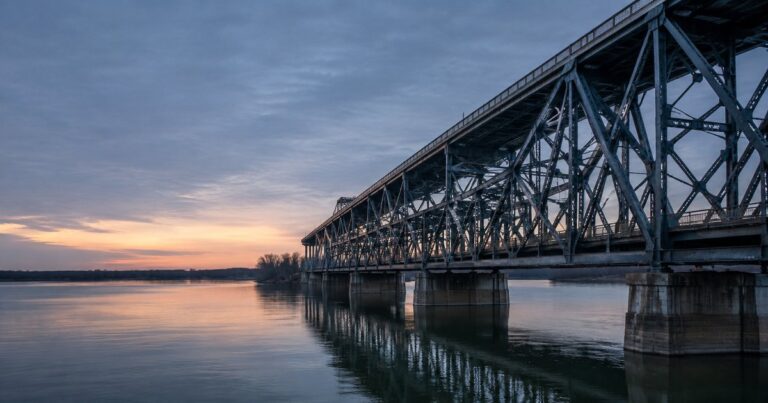 A weathered steel double-deck truss bridge crossing a wide Midwestern river at dusk, its lower roadway deck tracing the crossing directly while the upper deck runs the same line but extends past the lower deck onto a higher approach span on the far bank, in a cool gray and blue-teal industrial palette with no people present — a visual metaphor for the difference between a follow-form excess layer that stops where the primary stops and a commercial umbrella layer that reaches farther.