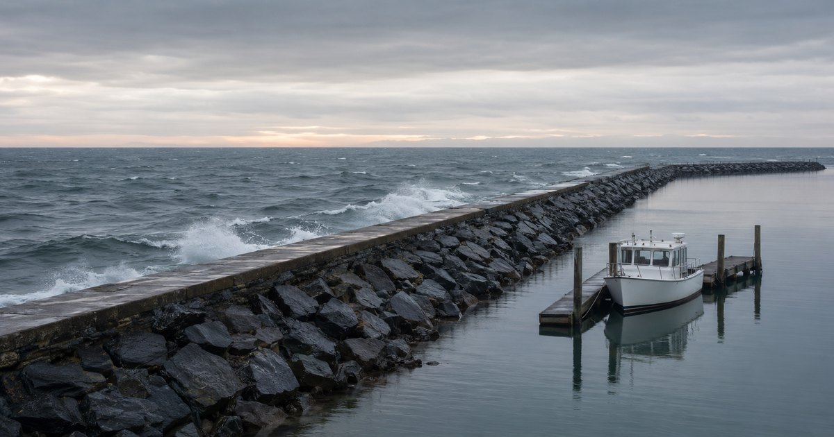 A long rubble-mound stone breakwater with a concrete cap at overcast dawn, steel-gray waves breaking white against its outer face while the harbor water inside lies calm and glassy, a single small white workboat moored level at a timber dock in the sheltered water with no people present — a visual metaphor for corporate indemnification and Side A D&O coverage as the layered wall a director's personal assets shelter behind.